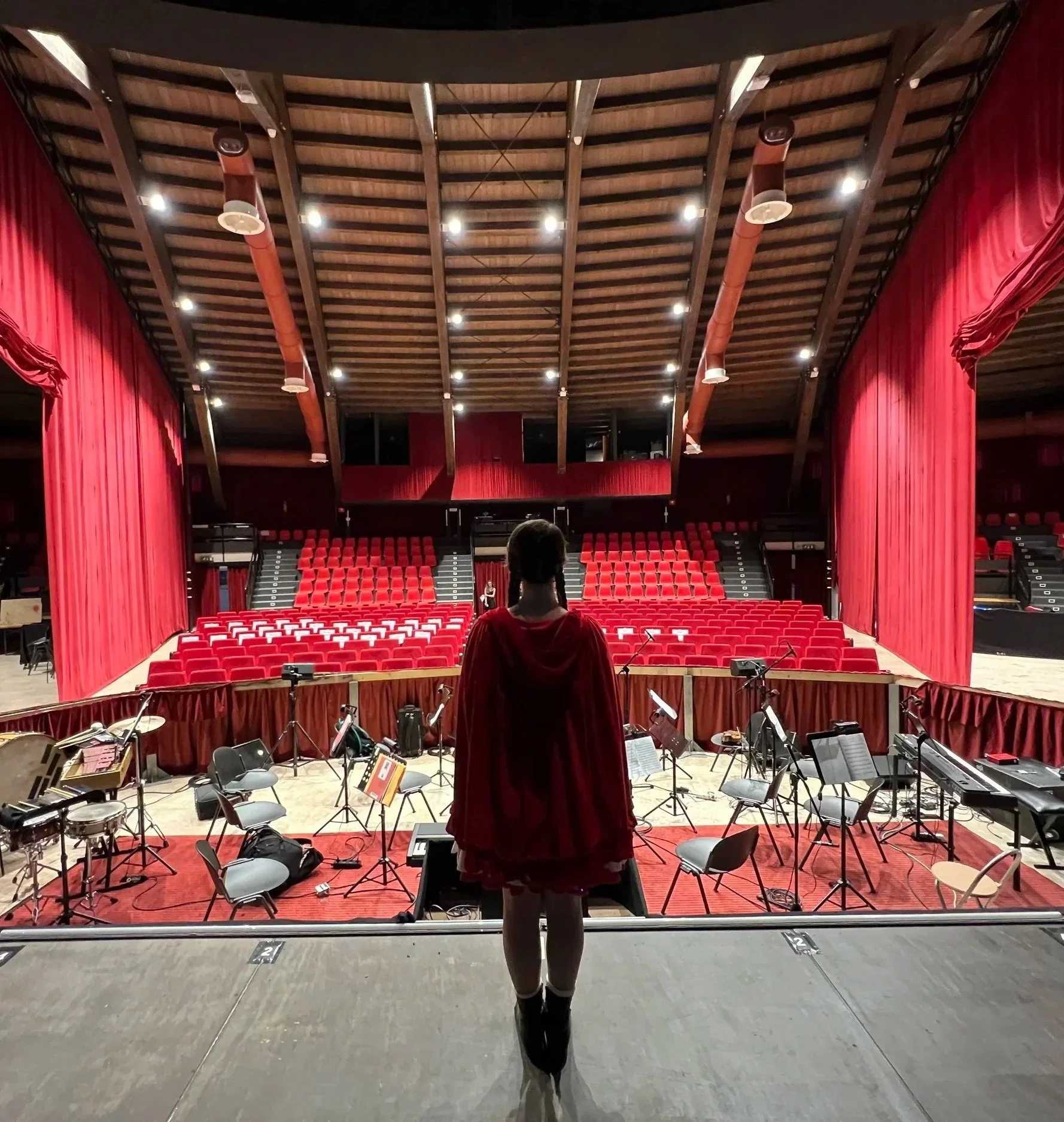 A girl stands on a stage, facing an auditorium with red seating, surrounded by musical instruments and music stands, preparing for a performance.