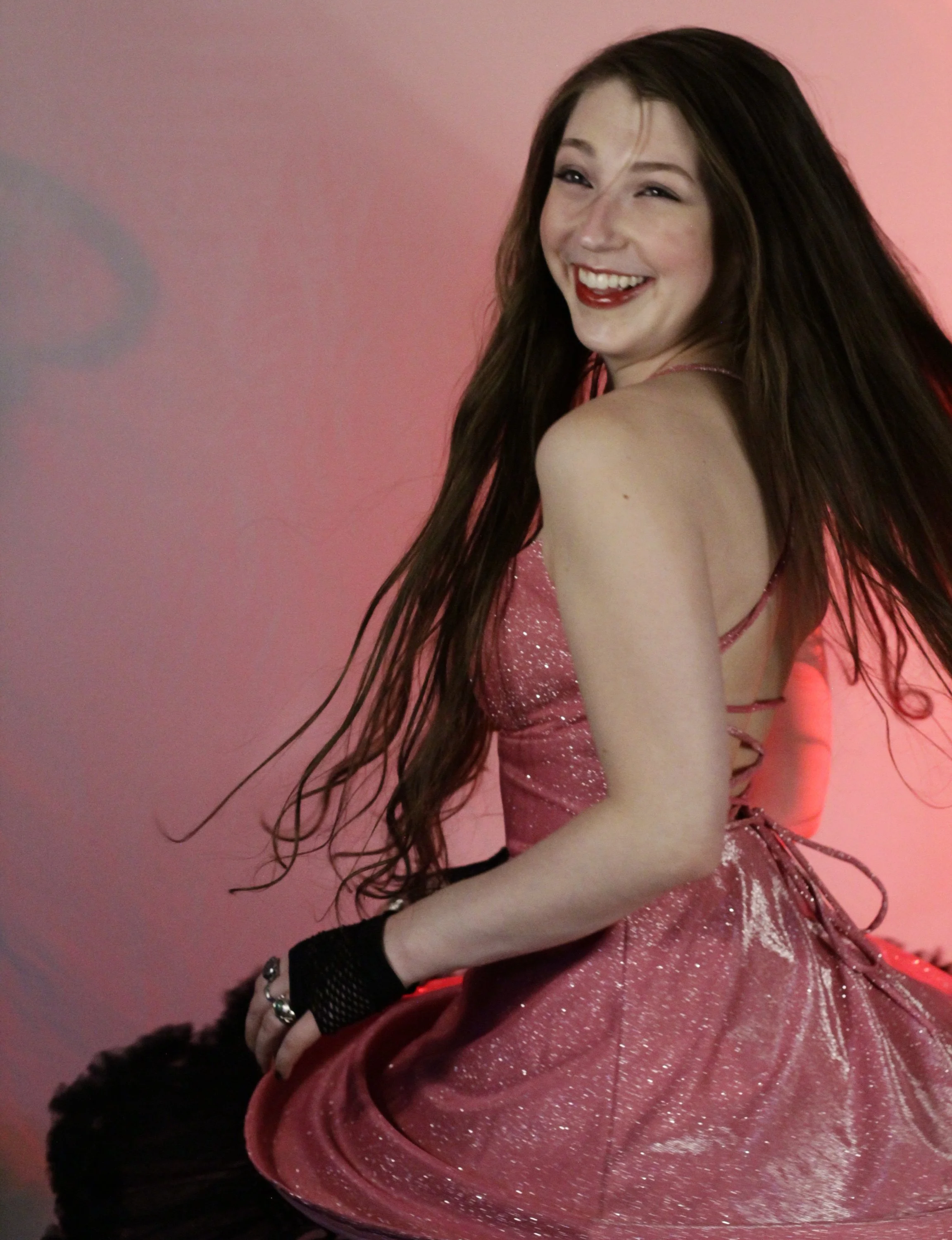 A young woman with long brown hair, wearing a pink sparkly dress and black gloves, sitting and smiling against a pink background.