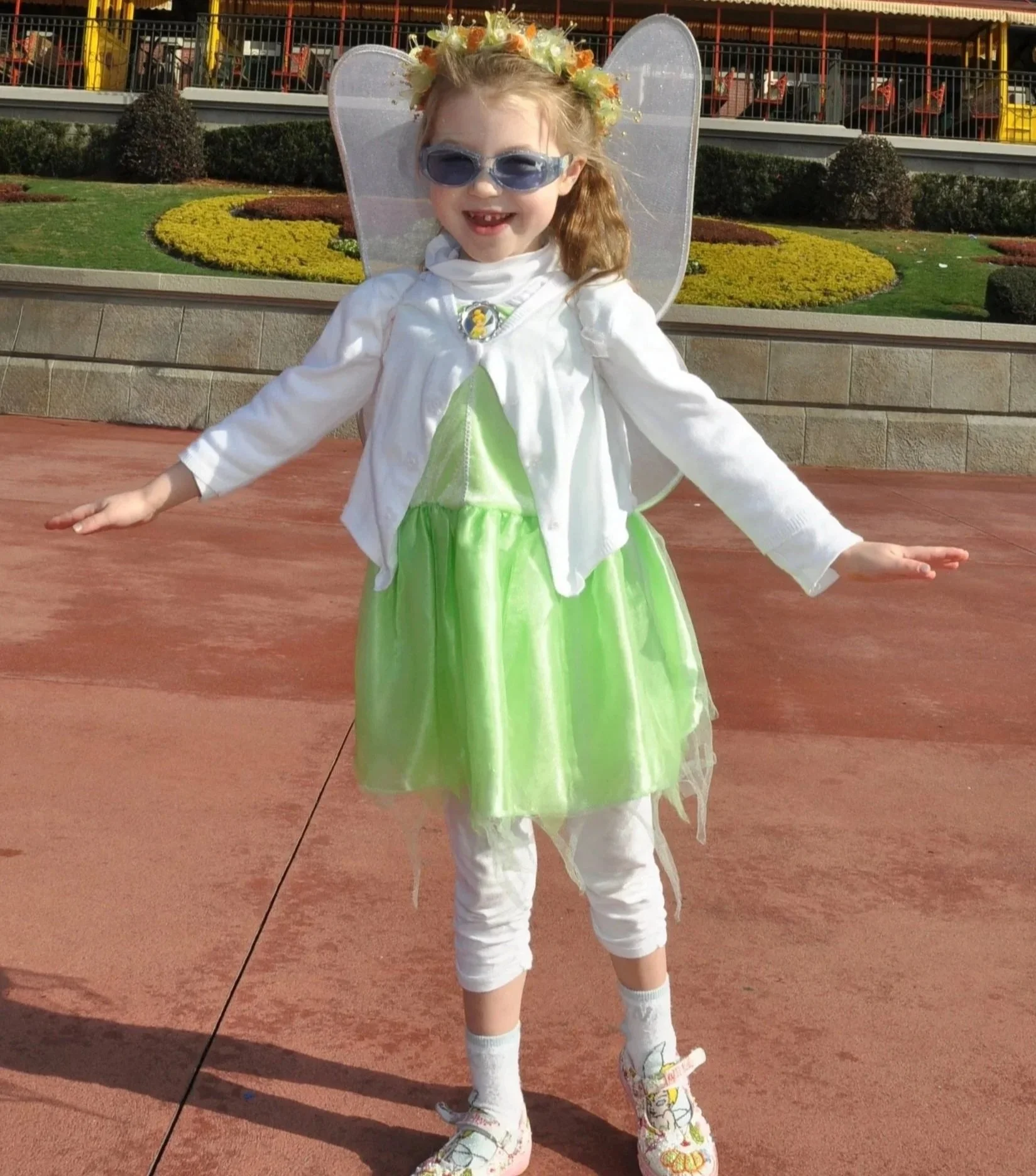 A young girl dressed as a fairy or princess, wearing a green dress, white jacket, fairy wings, fairy antennae headband, sunglasses, and colorful sneakers, standing outdoors with her arms outstretched, smiling.