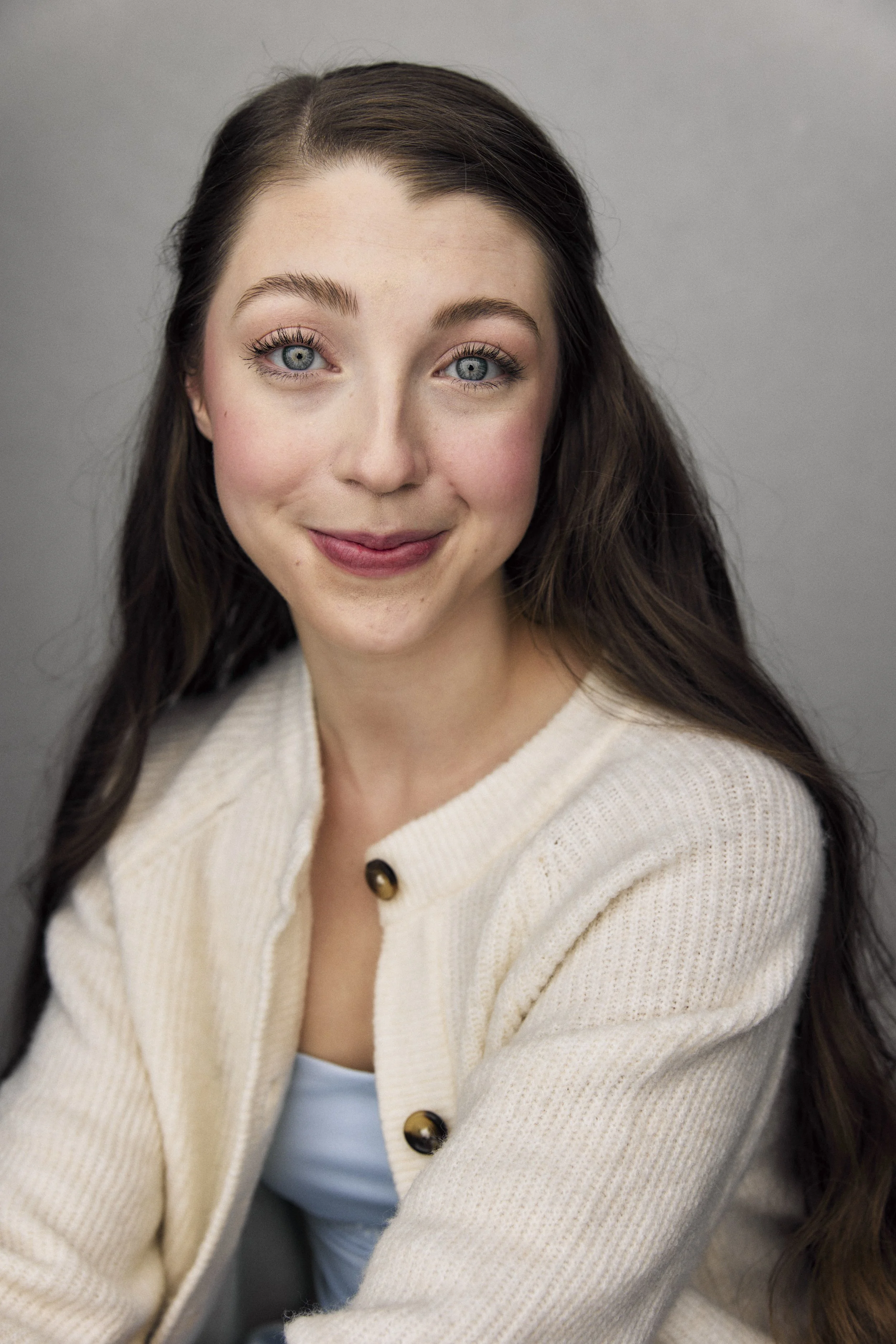 Close-up of a woman with long dark hair, blue eyes, wearing a cream-colored cardigan with dark buttons, smiling softly against a plain gray background.