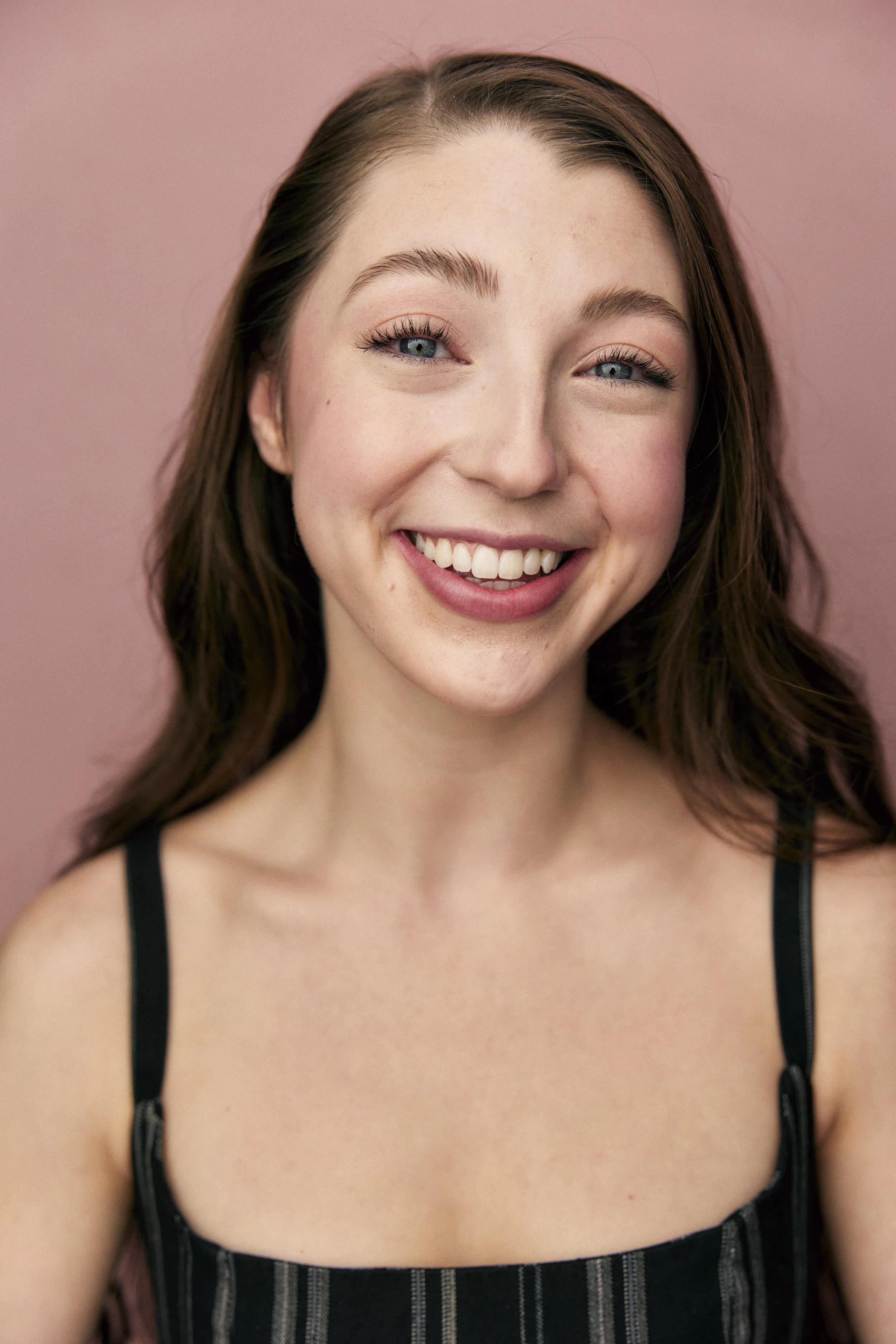 Close-up of a young woman with long brown hair, blue eyes, and a bright smile, wearing a black striped top, standing against a pink background.