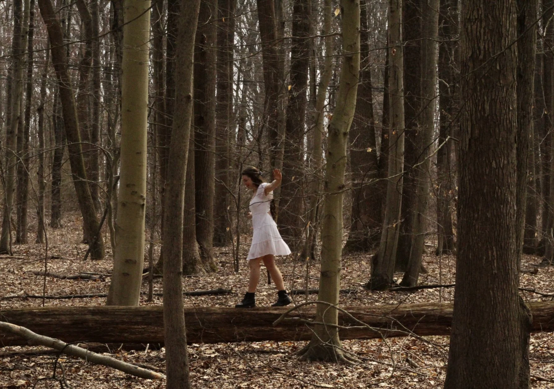 A woman in a white dress and black boots walking on a fallen tree in a wooded forest.
