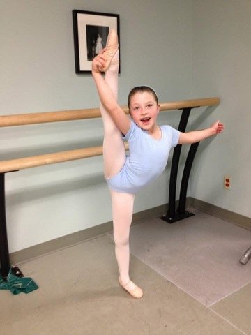 A young girl in a ballet studio performing an extended leg stretch at the barre, smiling.