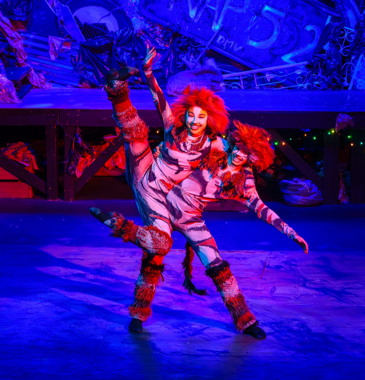 Two performers dressed in colorful, animal-print costumes with furry accents are dancing on stage under vibrant blue and red lighting, smiling and striking energetic poses.