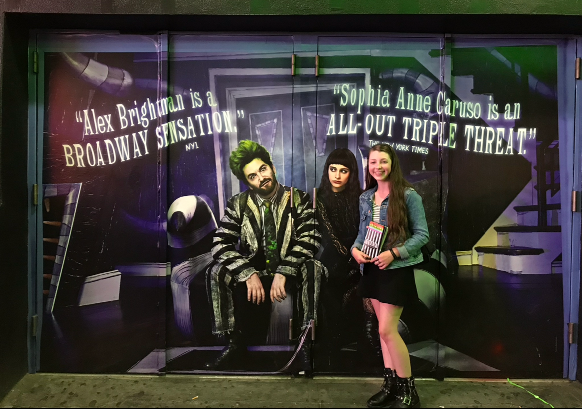 A young girl with long brown hair, wearing strips and combat boots, standing next to a large photo of two actors from the musical 'Beetlejuice.' The photo features the two actors in character, with quotes from reviews above them.