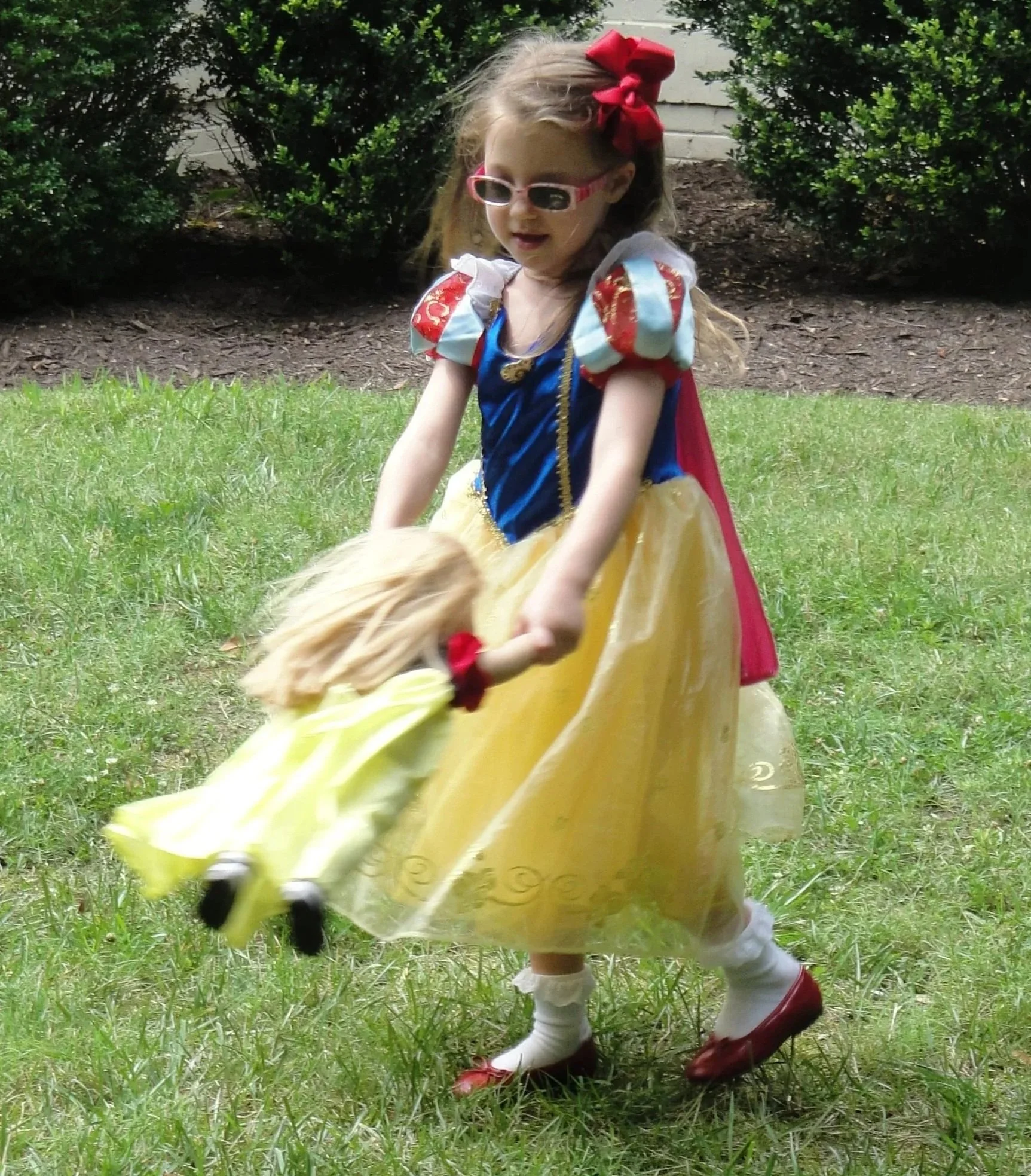 A young girl dressed as Snow White holding a blonde doll outdoors on grass with bushes in the background.