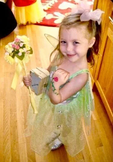 A young girl dressed as a fairy holding a bouquet of flowers and a gift bag, smiling indoors on a wooden floor.