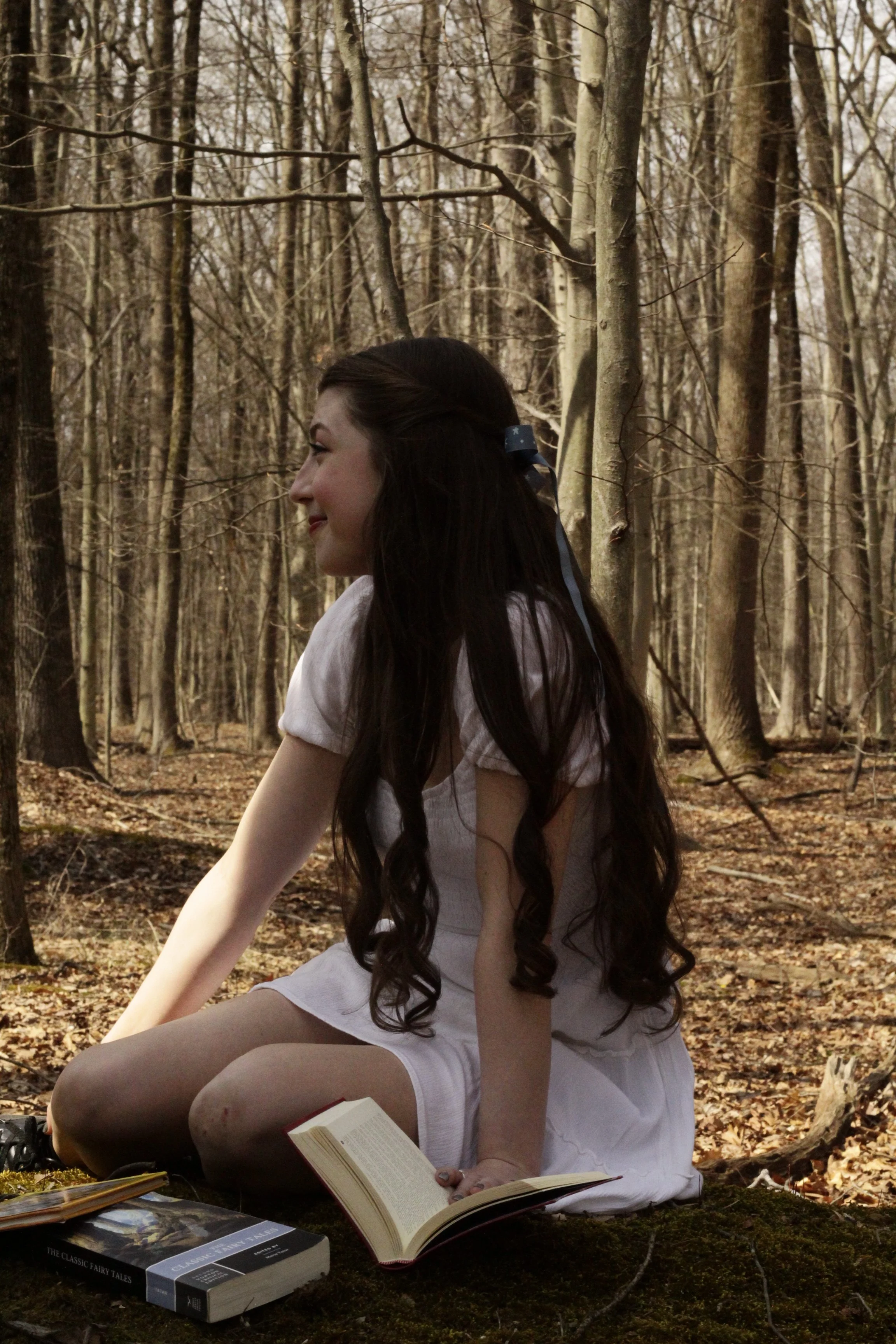 A young woman with long dark hair sitting on mossy ground in a forest, reading a book, with several other books nearby, wearing a white dress.