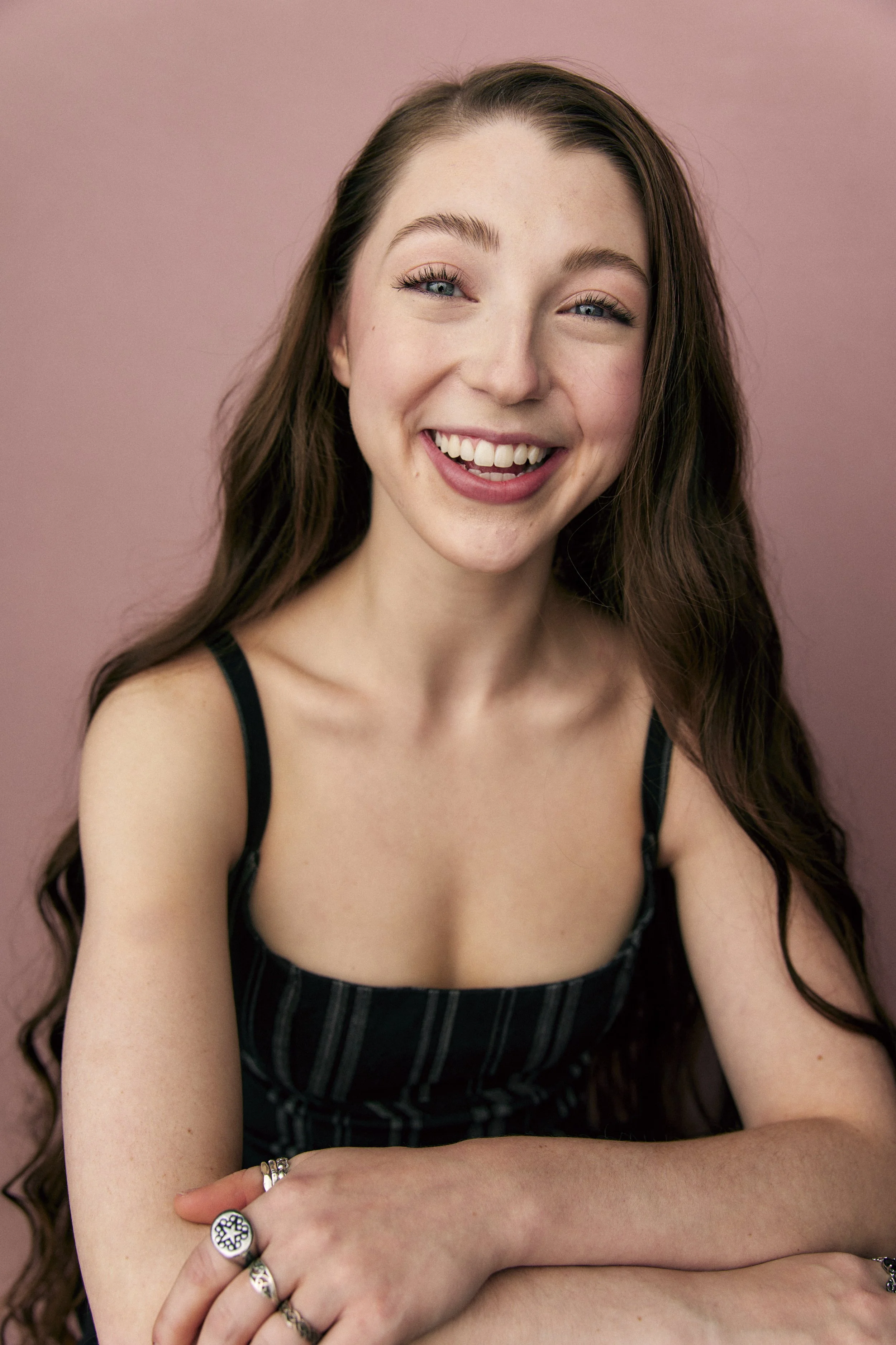 A young woman with long, wavy brown hair smiling at the camera against a pink background.