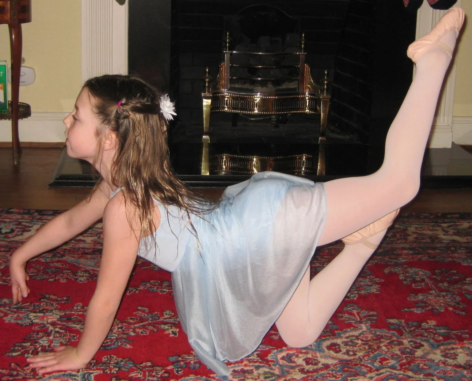 A young girl wearing a light blue leotard and ballet tights, posed on her hands and knees with her legs raised behind her, on a patterned rug in front of a fireplace.