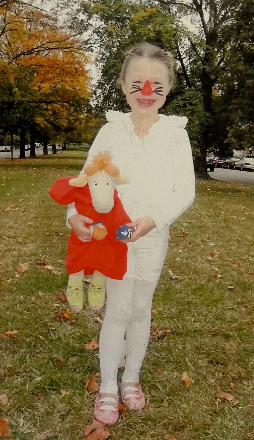 Person with face painted like a cat, holding a Giraffe stuffed animal, standing outdoors on grass with autumn leaves and trees in the background.
