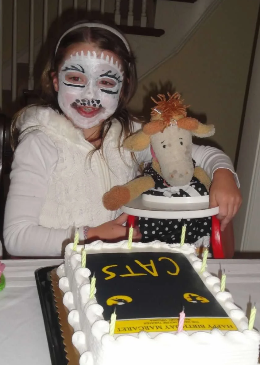 A girl with face paint resembling a cat, smiling and holding a stuffed animal of a Giraffe. There is a birthday cake with candles and a sign that reads 'CATS' in yellow letters.