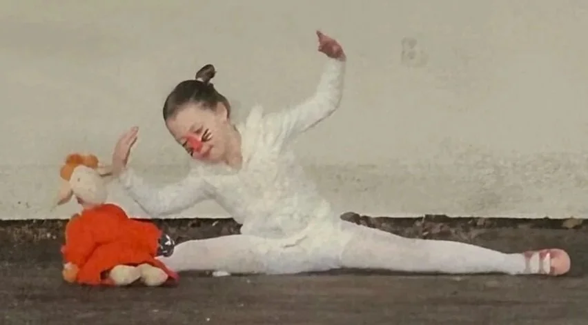 Young girl dressed as a white cats with face paint, stretching in a split on the floor. She is holding a stuffed giraffe doll and smiling.
