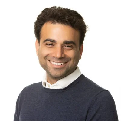 A smiling man with dark hair, wearing a dark sweater and white shirt, posing against a white background.