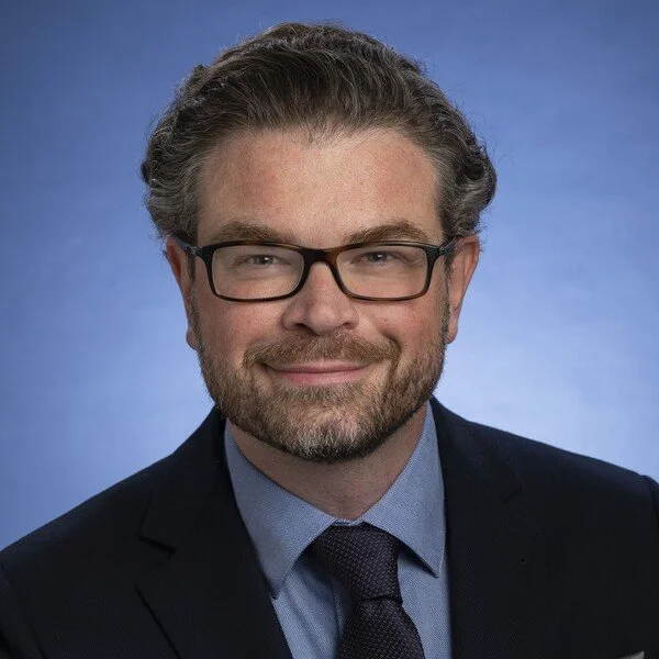 Professional headshot of a man with glasses, a beard, and gray hair, wearing a dark suit, blue shirt, and dark tie against a blue gradient background.