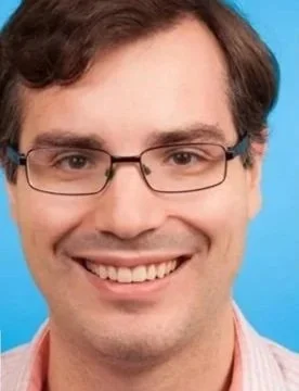 Close-up of a man with brown hair, glasses, and a big smile, wearing a light pink shirt, against a blue background.
