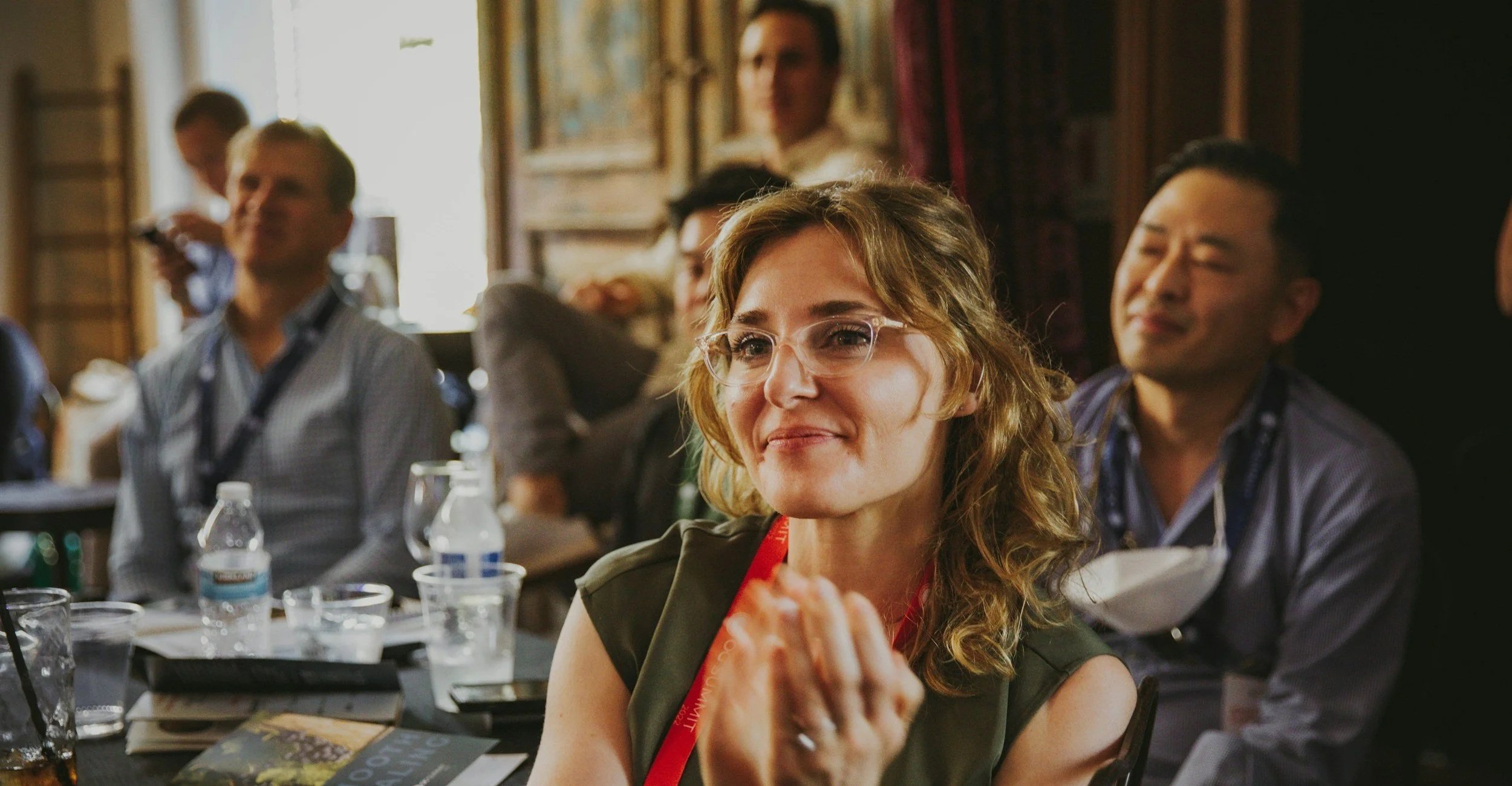 Group of people sitting at a conference or meeting, focused on a woman with curly blonde hair in the foreground, wearing glasses and a dark green shirt, with attentive expressions.