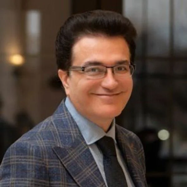 A man with dark hair, glasses, and a checked blazer smiling at the camera indoors.