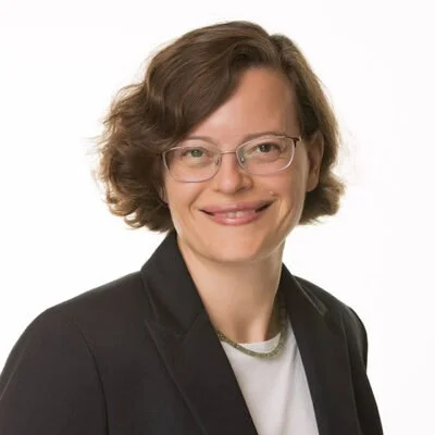 A woman with short, wavy brown hair wearing glasses, a black blazer, and a white shirt smiling against a plain white background.