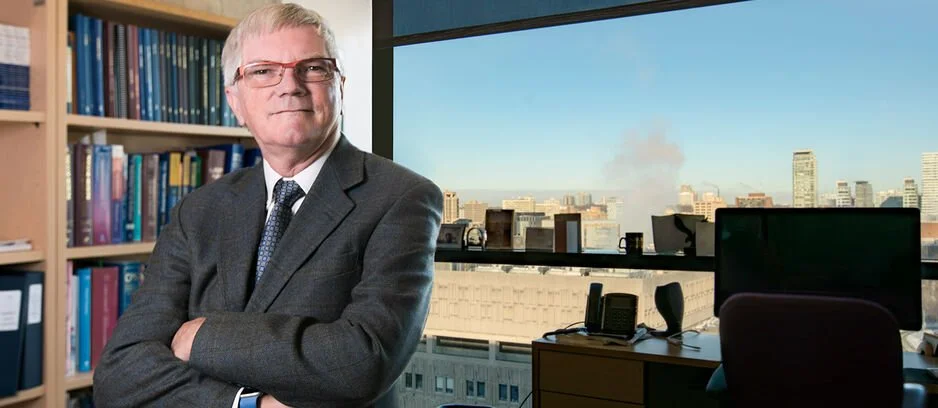 A professional man standing in an office with a city skyline view, bookshelves, and office equipment in the background.
