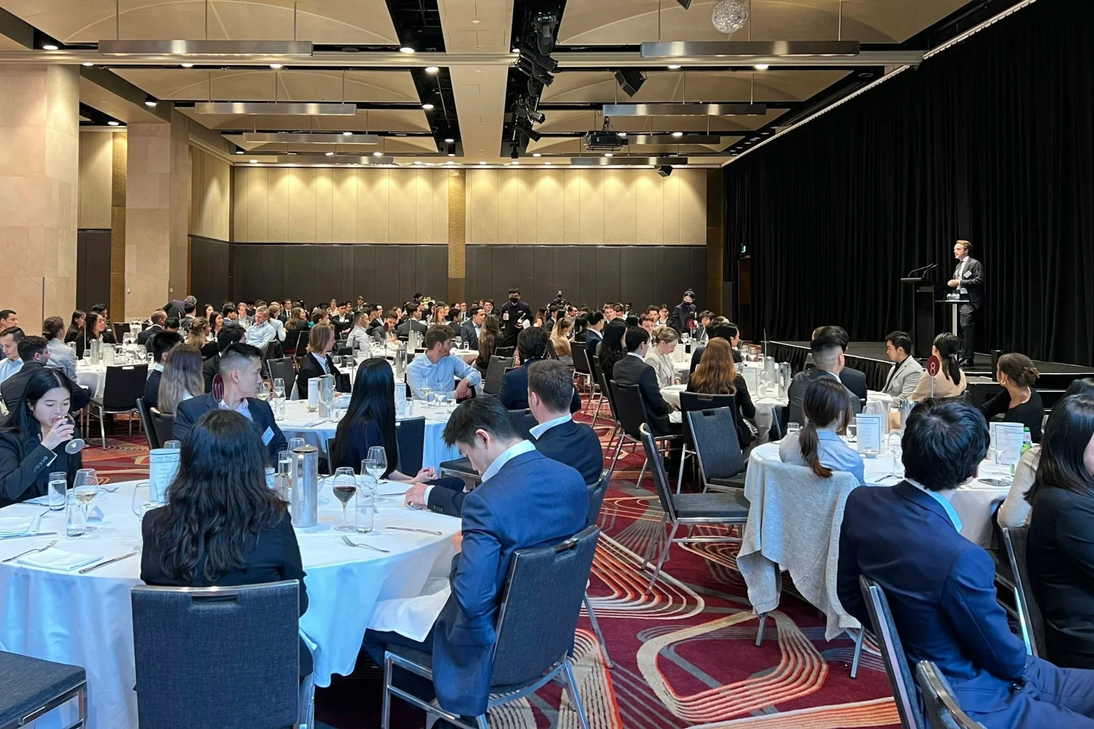 A large conference room filled with round tables of attendees, some eating and drinking, facing a stage where a speaker stands at a podium.