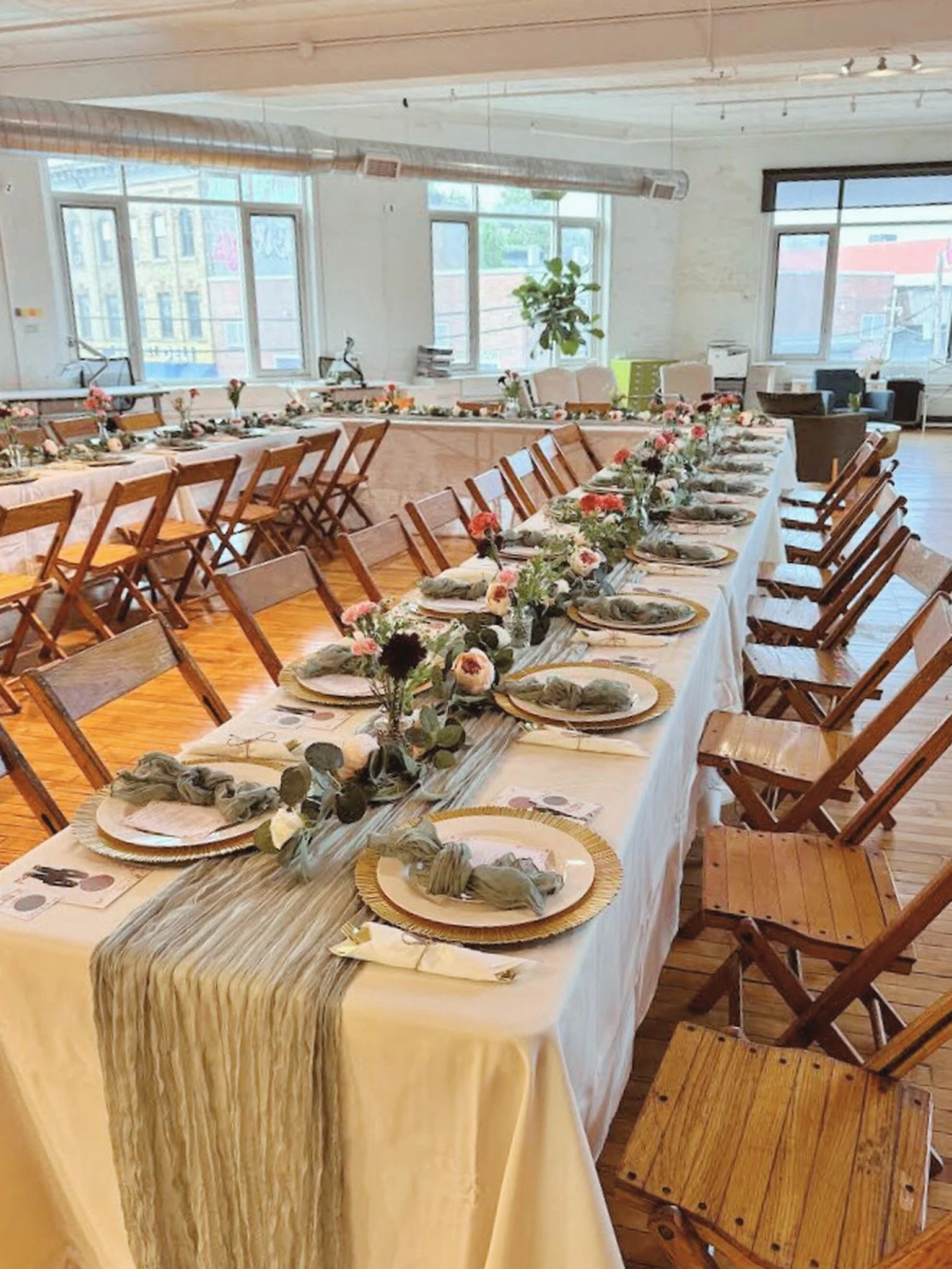 A long dining table set for a celebration in a bright, modern room with large windows. The table has a white tablecloth, greenery, and pink and white flowers as a centerpiece. Each place setting includes a gold charger plate, a napkin, and printed me