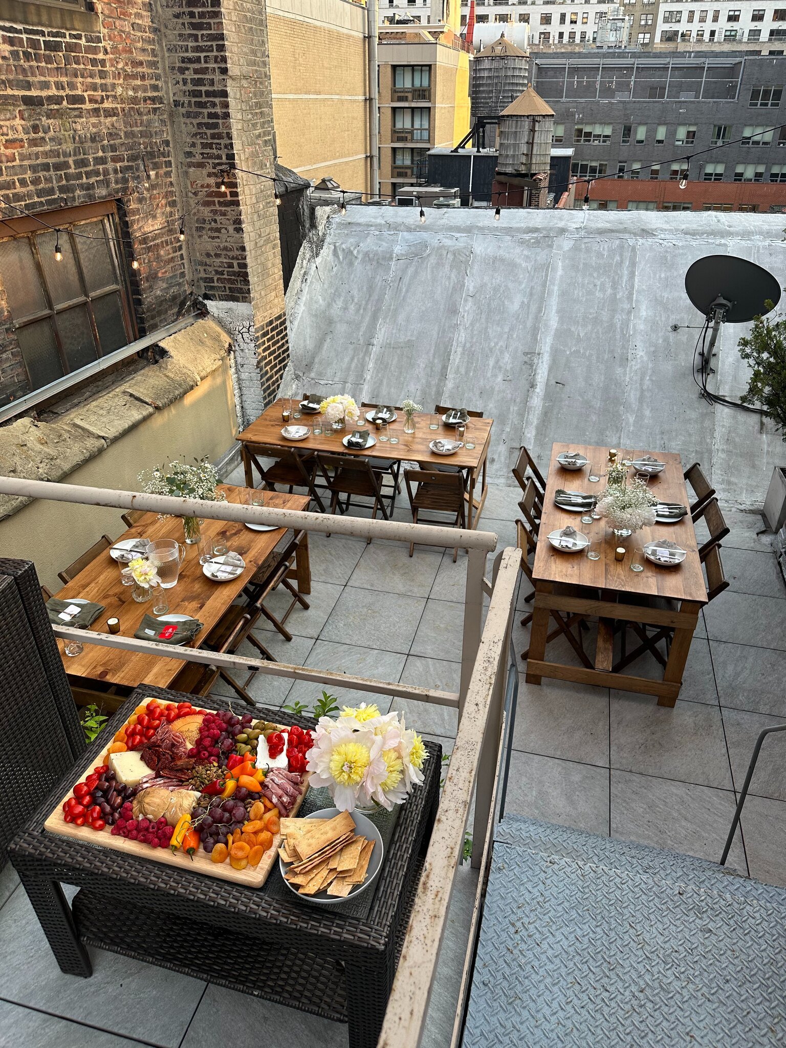 Rooftop dining area with three wooden tables set with black napkins, white plates, and glasses. The tables are decorated with white flowers and small candles. A black wicker table in the foreground is topped with a charcuterie board featuring cheese,