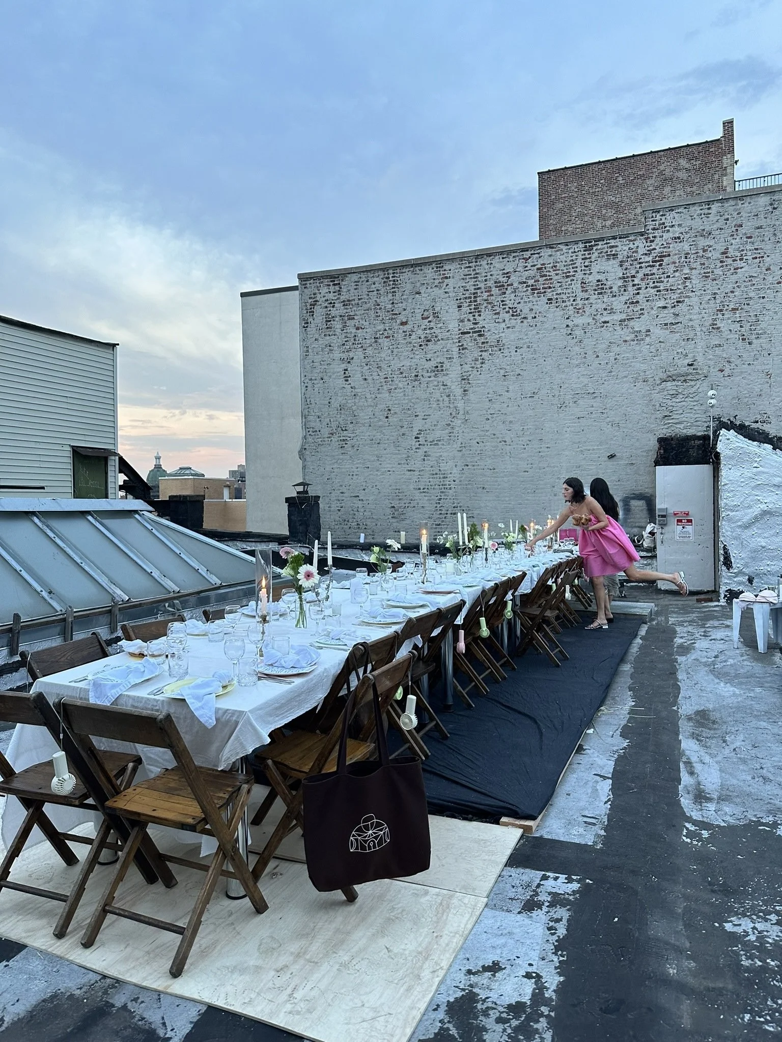 Rooftop outdoor dinner setup with a long table decorated with candles, flowers, and tableware, with two women in pink dresses setting the table against a brick wall in the evening sky.