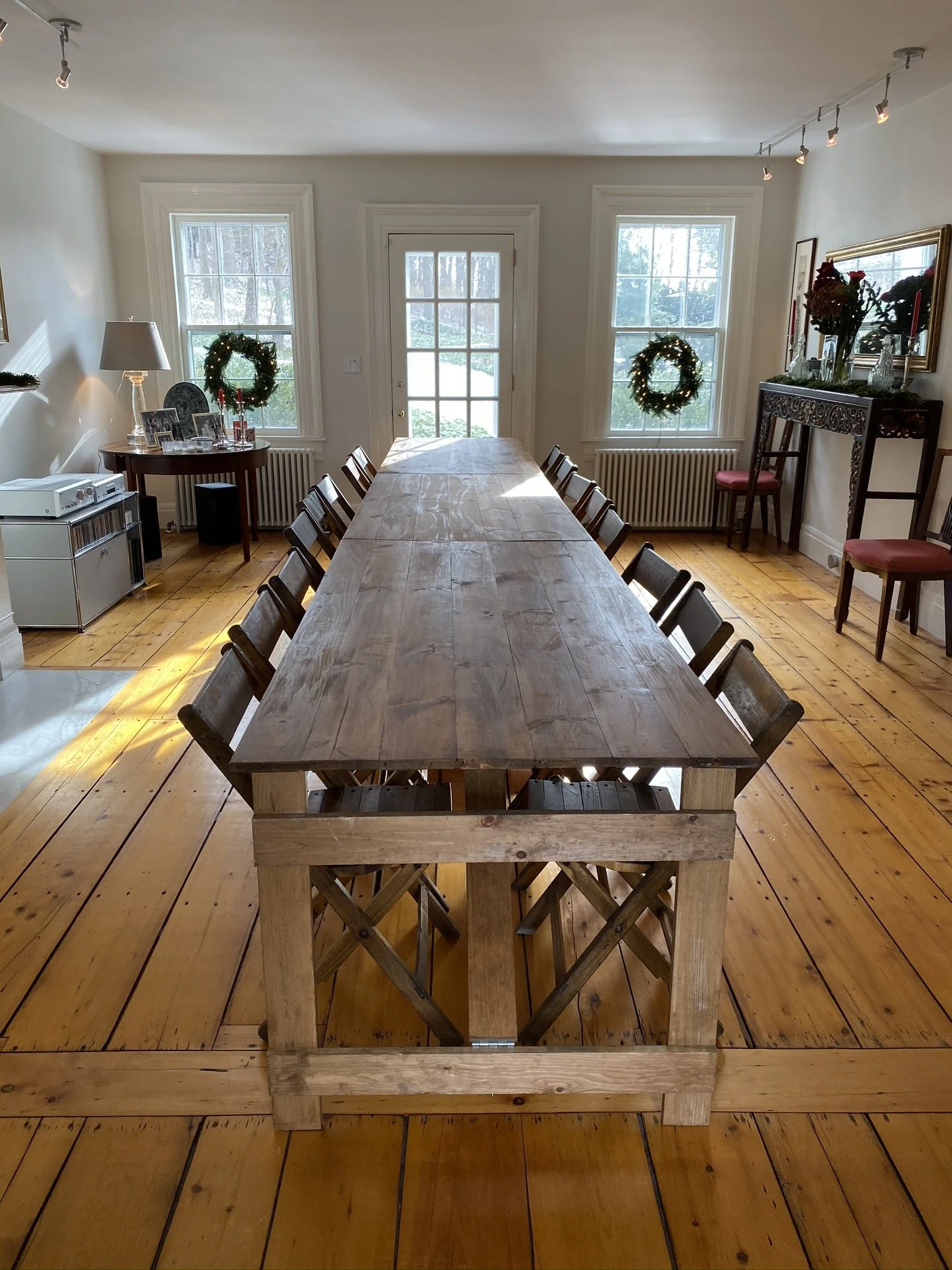 A long banquet of two handmade folding farm tables with vintage wooden folding chairs set of Christmas dinner in a country home dining room with windows, wreaths, and holiday decor.