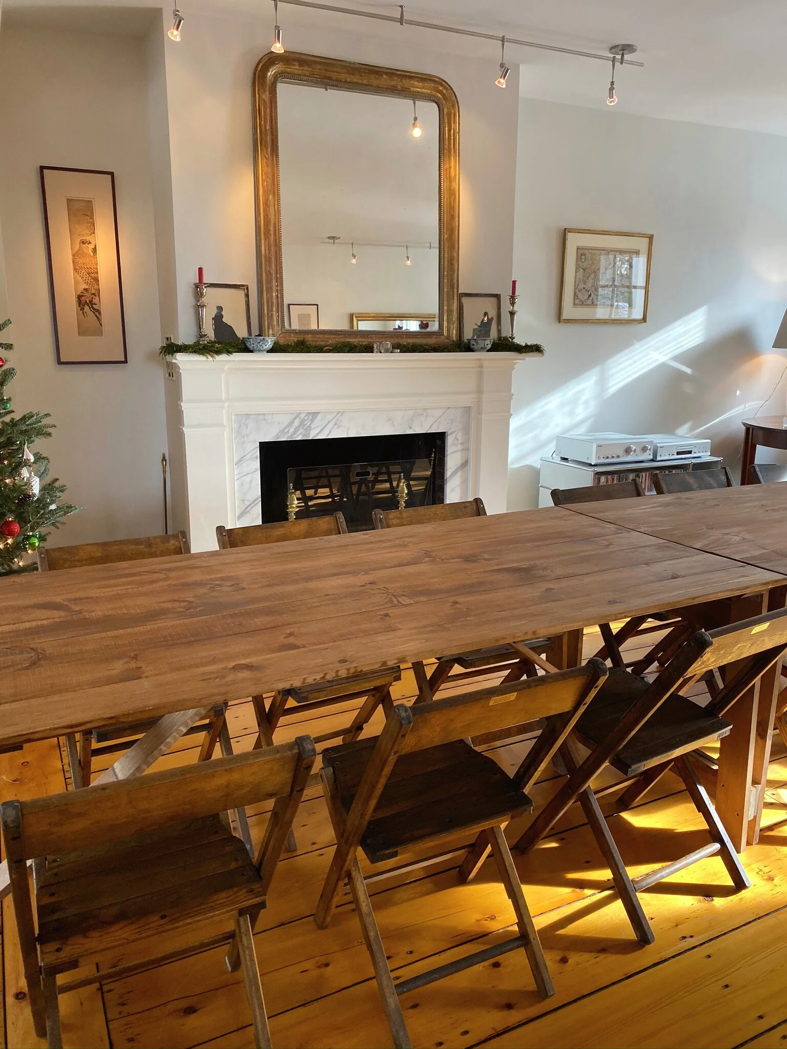 A dining room with a large wooden table, wooden chairs, a fireplace with a gold-framed mirror above, and holiday decorations on the mantle.