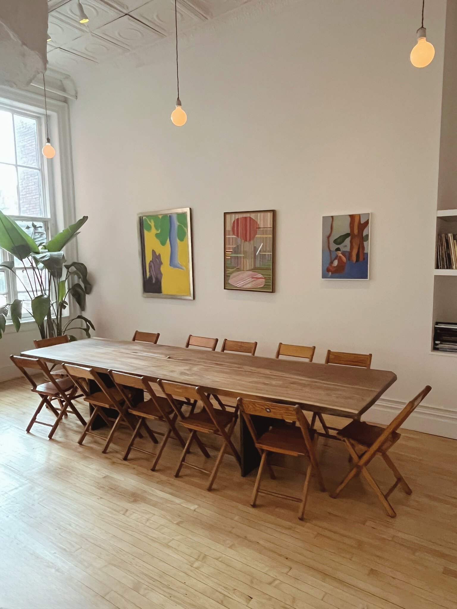 Interior of a room with a long wooden table and multiple foldable chairs. There are three colorful abstract paintings on the white wall, a large window with a plant nearby, pendant lights hanging from the ceiling, and wooden flooring.