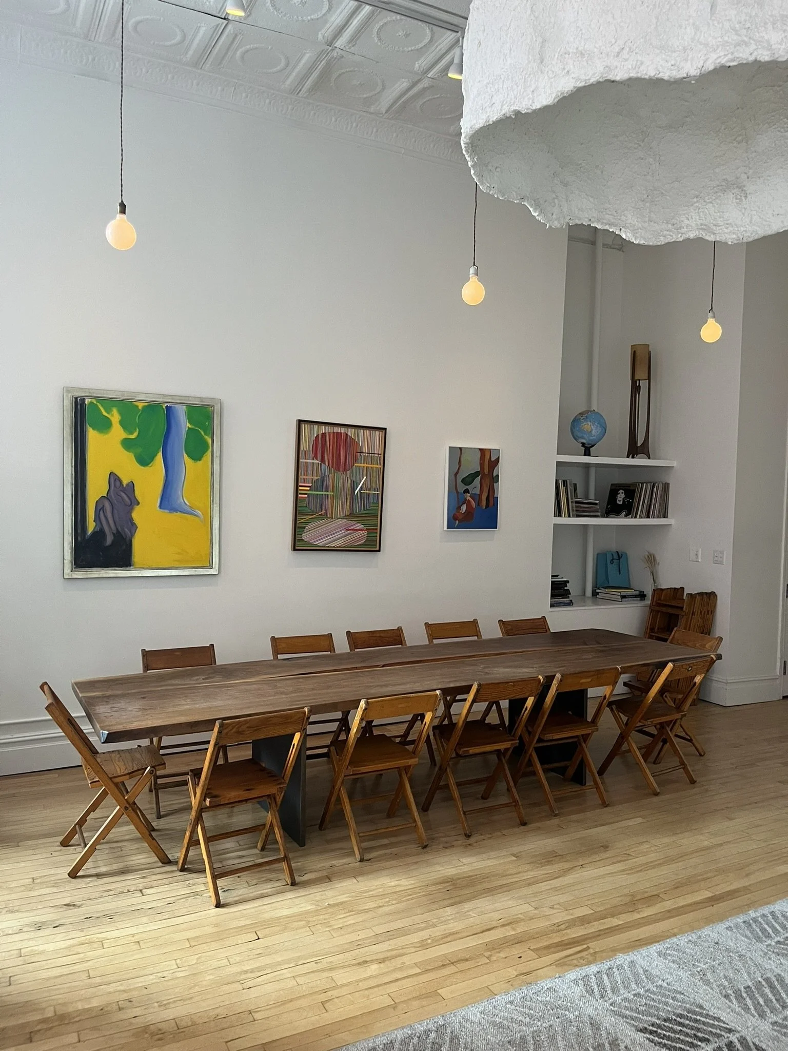 A long wooden dining table with 12 wooden folding chairs in a room with light hardwood floors, white walls, and colorful artwork hanging on the wall. There are three exposed light bulbs hanging from the ceiling and a distinctive textured, cloud-like 