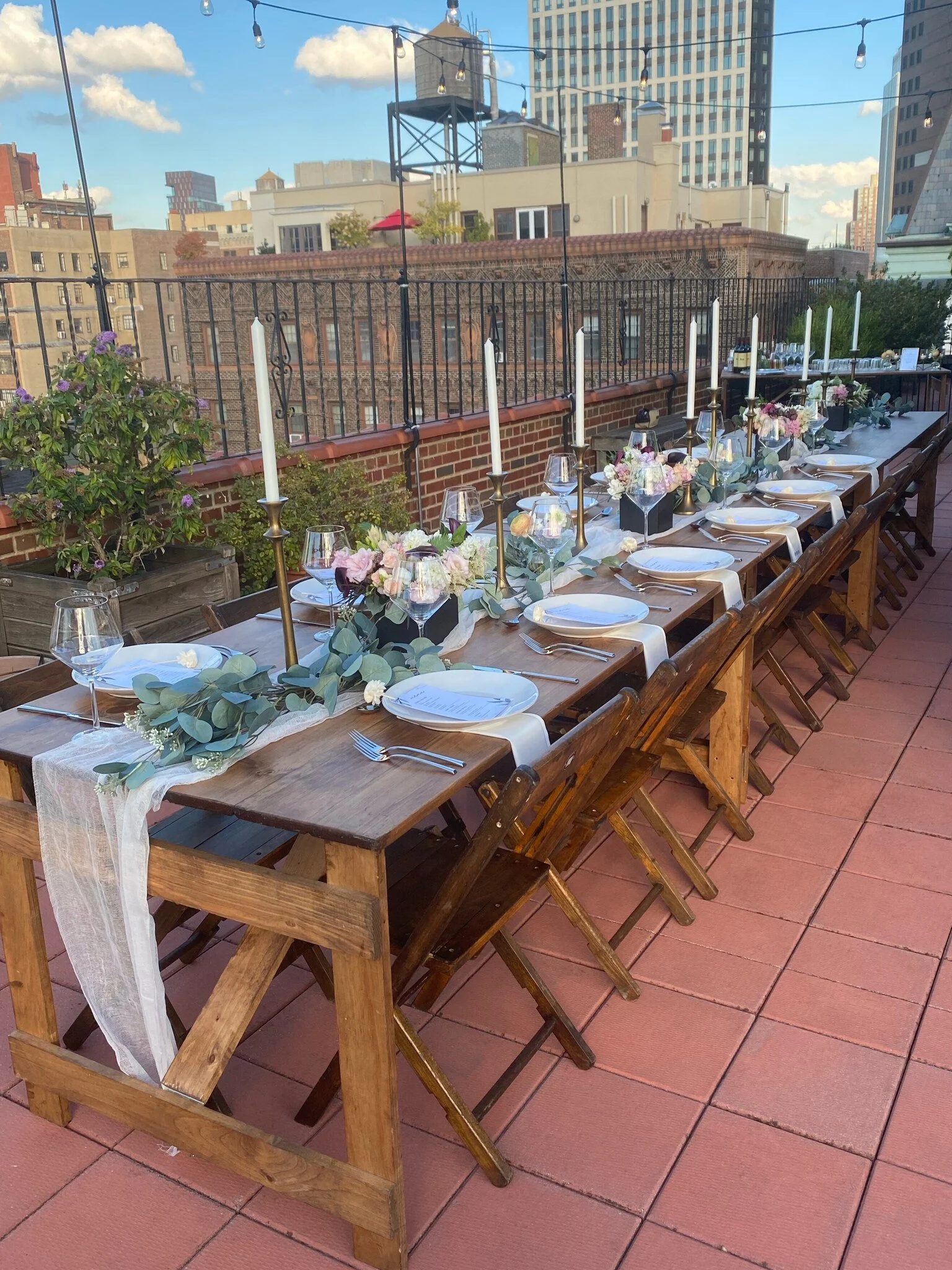 A banquet of handmade folding farm tables with vintage wooden folding chairs set and decorated for a birthday lunch on a Brooklyn Heights rooftop with views of blue sky and city buildings in the background.