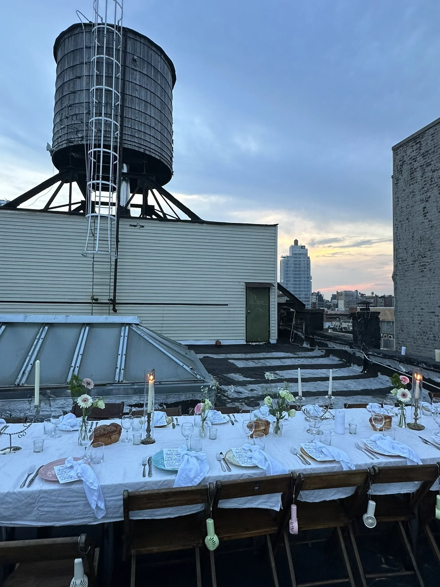 Vintage wooden folding chairs at a long table set for dinner at a rooftop party overlooking city buildings and a giant water tower at sunset.