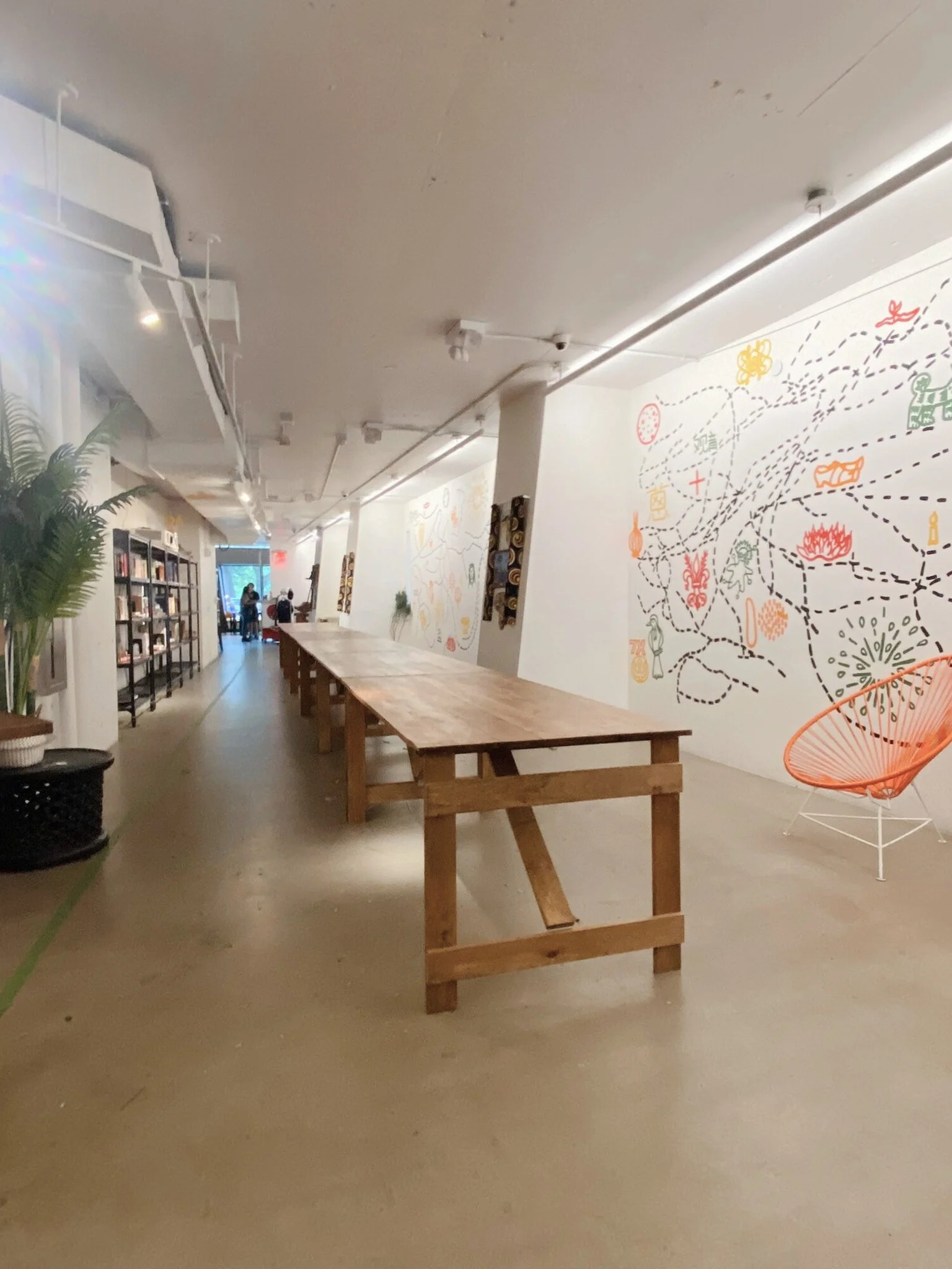 Interior of a modern art gallery or cafe with a long wooden table, potted plants, bookshelves, colorful wall mural, orange chair, and people in the background.