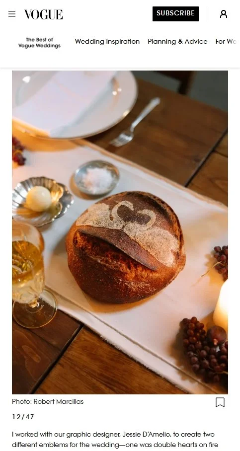 A round loaf of bread with a design of a heart shaped emblem on top, placed on a white table runner. Surrounding the bread are a glass of white wine, a small silver dish with a butter pat, and a plate with a butter knife. There are grapes and a lit candle on the table, with a cake or bread dish in the background.