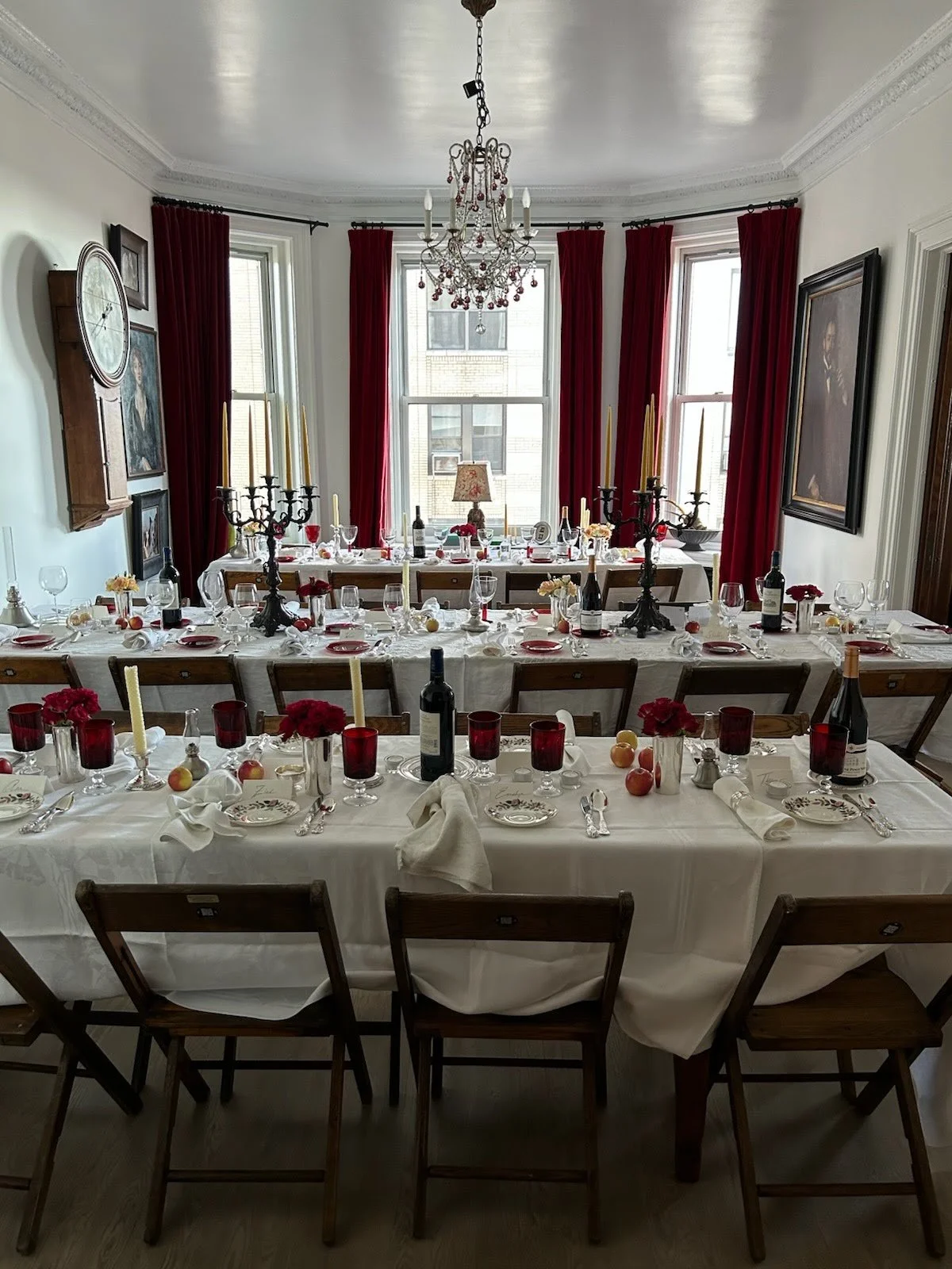 A dining room set for a formal dinner with a long table, red candles, wine bottles, wine glasses, floral centerpieces, and red and white tableware. Red curtains hang in front of large windows, and a chandelier hangs from the ceiling.