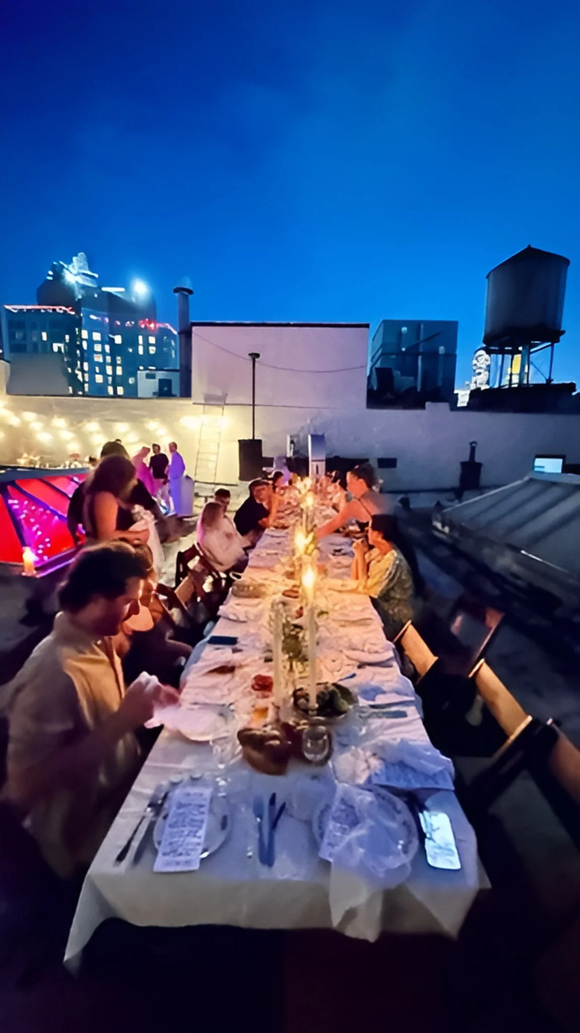 Long dining table with people seated in vintage wooden folding chairs,  eating food, lit by candles on a Manhattan rooftop during twilight, with city buildings and a large water tower in the background.
