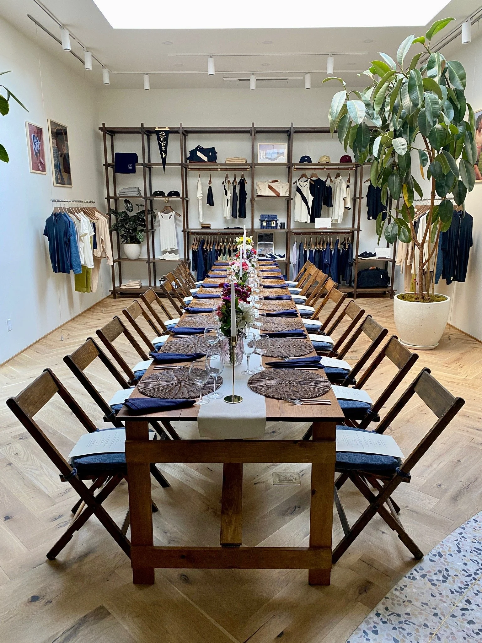 A dining table set with plates, napkins, glasses, and flowers in a retail clothing store, with shelves of clothing and accessories in the background.