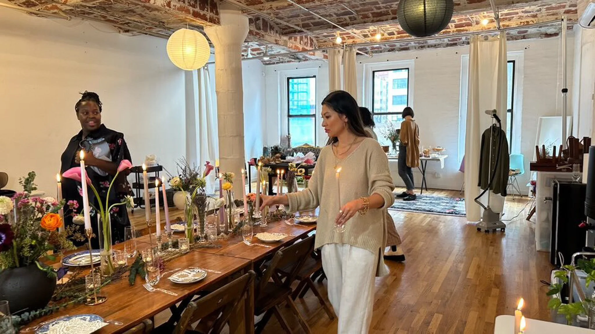 Women preparing a decorated table with candles and flowers in a bright, modern room.
