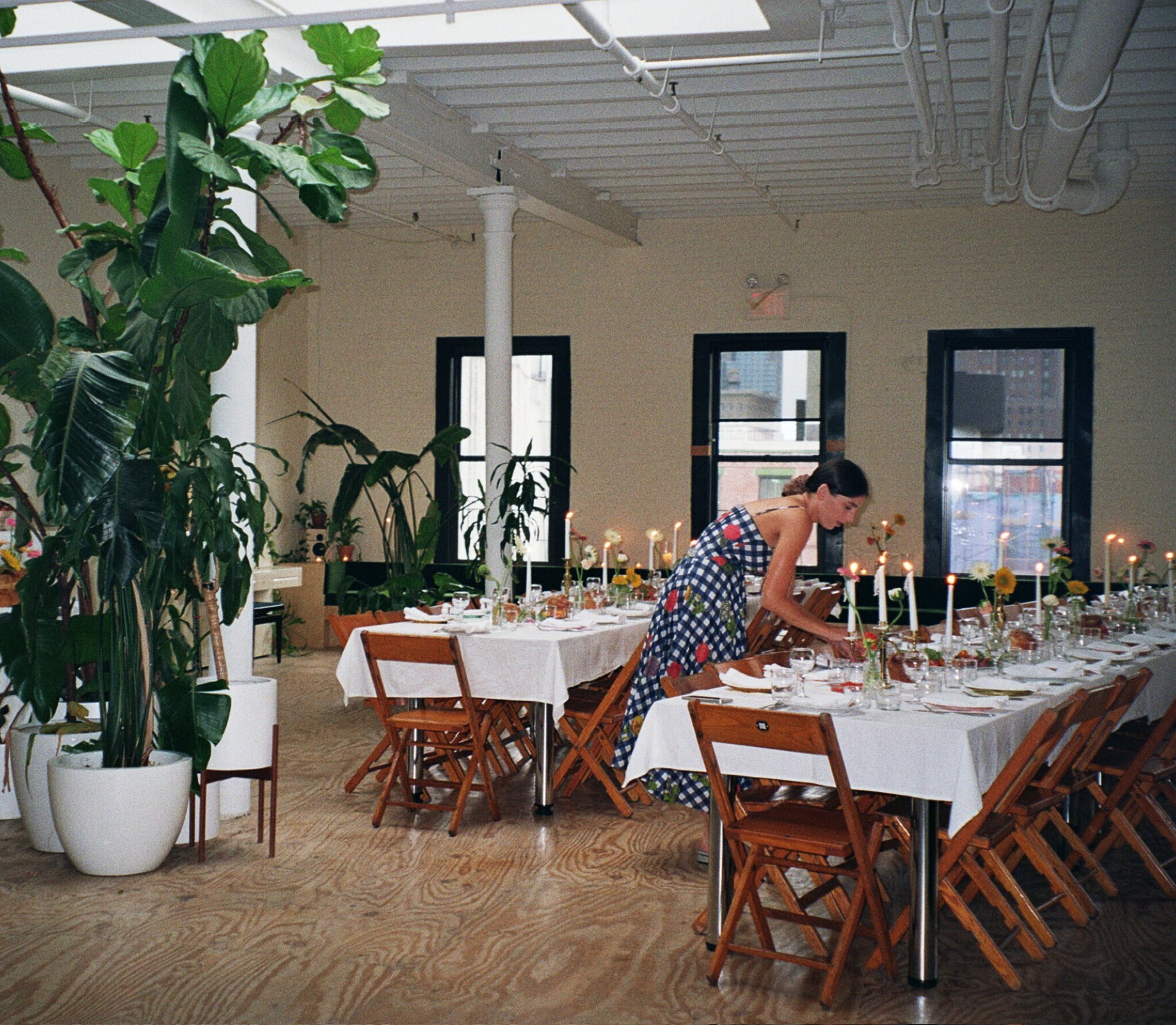 Woman lighting candles on a long banquet table surrounded by vintage wooden folding chairs in a Chinatown event space.