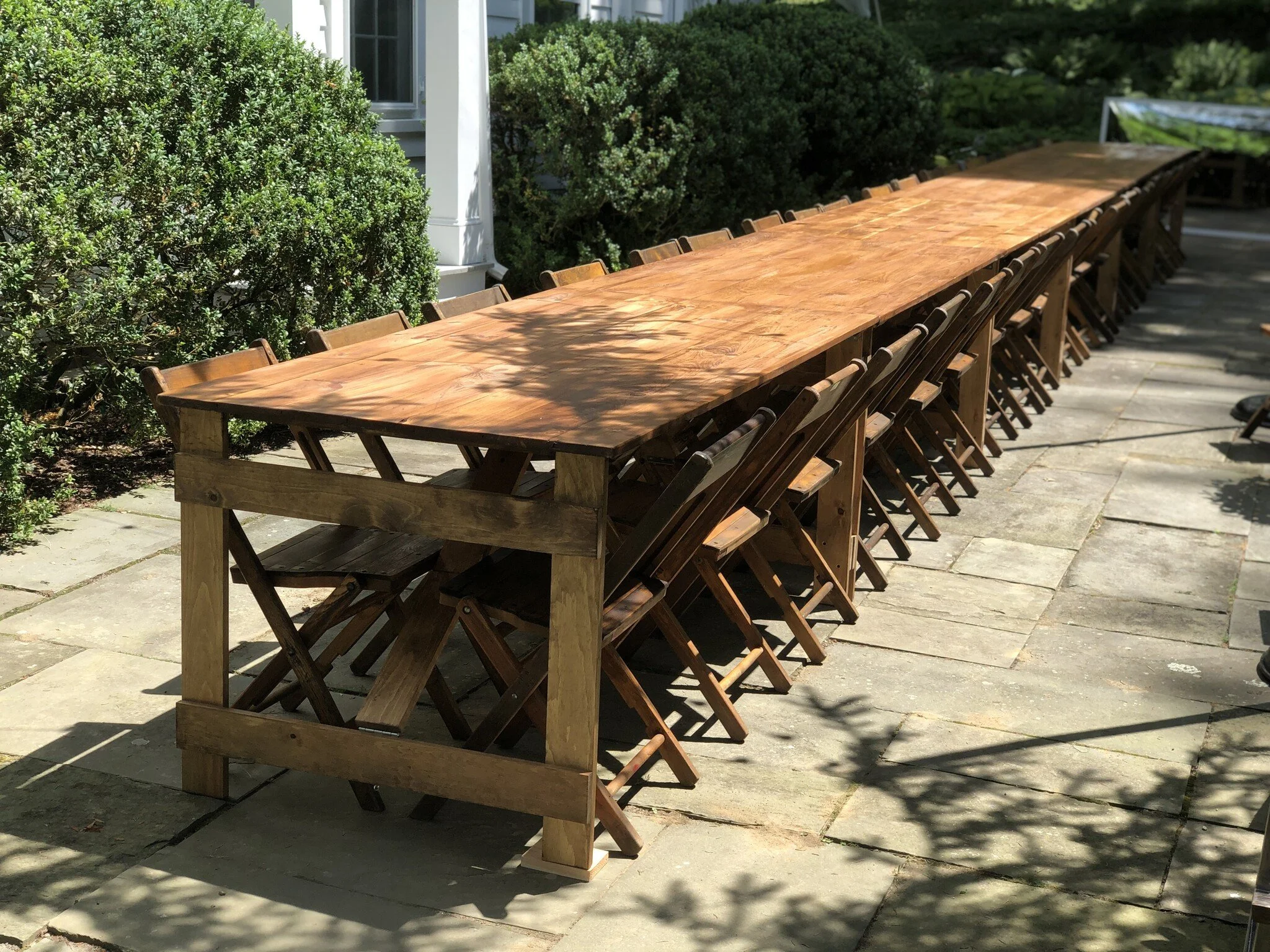 Long outdoor wooden dining table with folding chairs on a stone patio, surrounded by bushes.