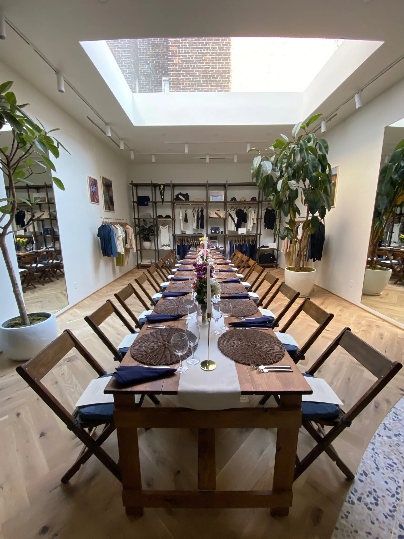 A large rectangular dining table set for a meal inside a bright room with a skylight. The table has woven placemats, white napkins, wine glasses, silverware, and floral centerpieces. The room is decorated with potted plants, mirrors, and clothing dis
