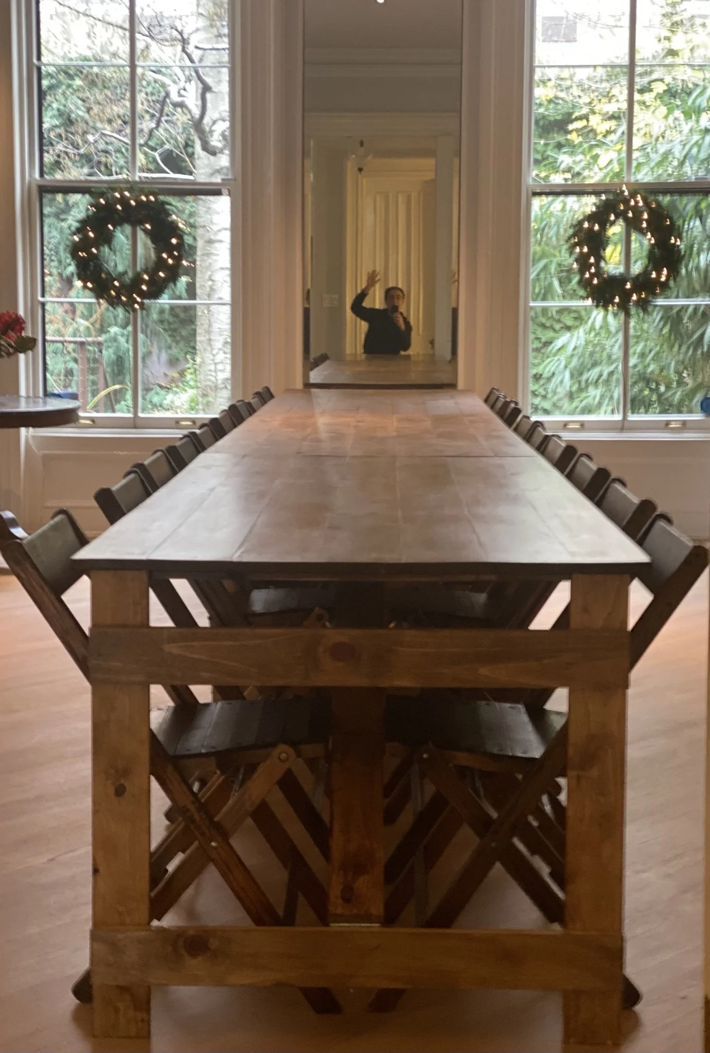 A long wooden dining table with chairs in a bright room with large windows and holiday wreaths, a person taking a photo in the mirror behind the table.