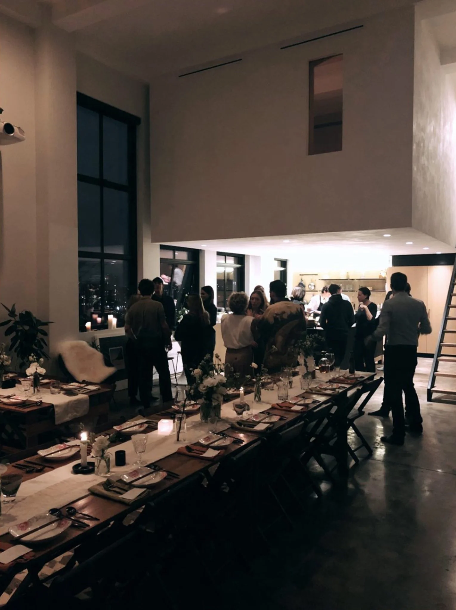 A long banquet of handmade folding farm tables and vintage wooden folding chairs at a dinner party in a dimly lit modern loft apartment.