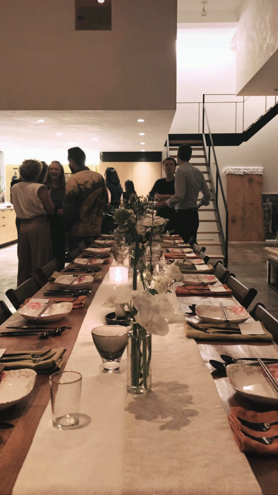 A banquet of handmade folding farm tables with vintage wooden folding chairs and people mingling and talking at a dinner party in a modern, well-lit home.