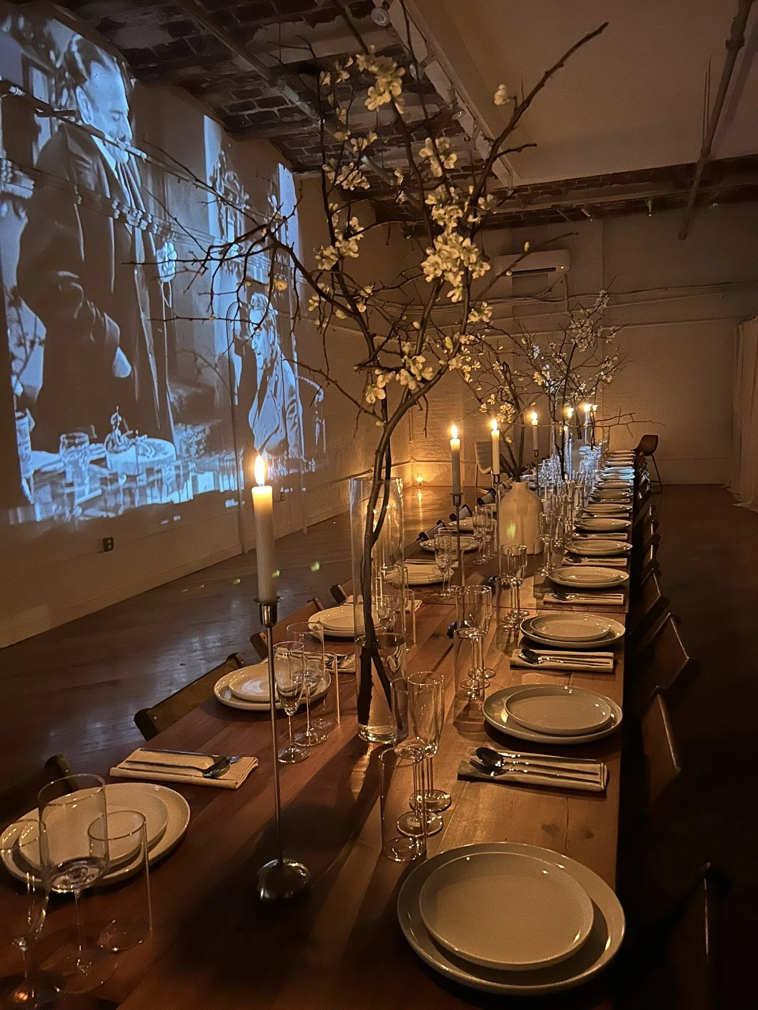 A long dining table decorated with vases of blossoming branches, surrounded by place settings, with lit candles and a vintage film projection of two people on the wall in the background.