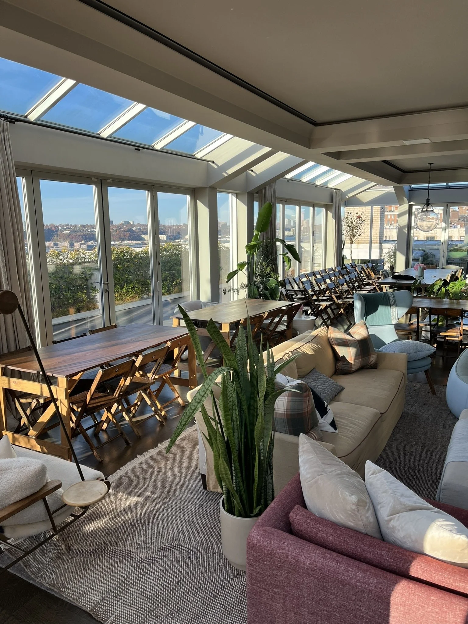 Interior of a sunlit room with large glass windows and a sloped glass ceiling, decorated with assorted sofas, wooden tables, and potted plants, overlooking a cityscape.