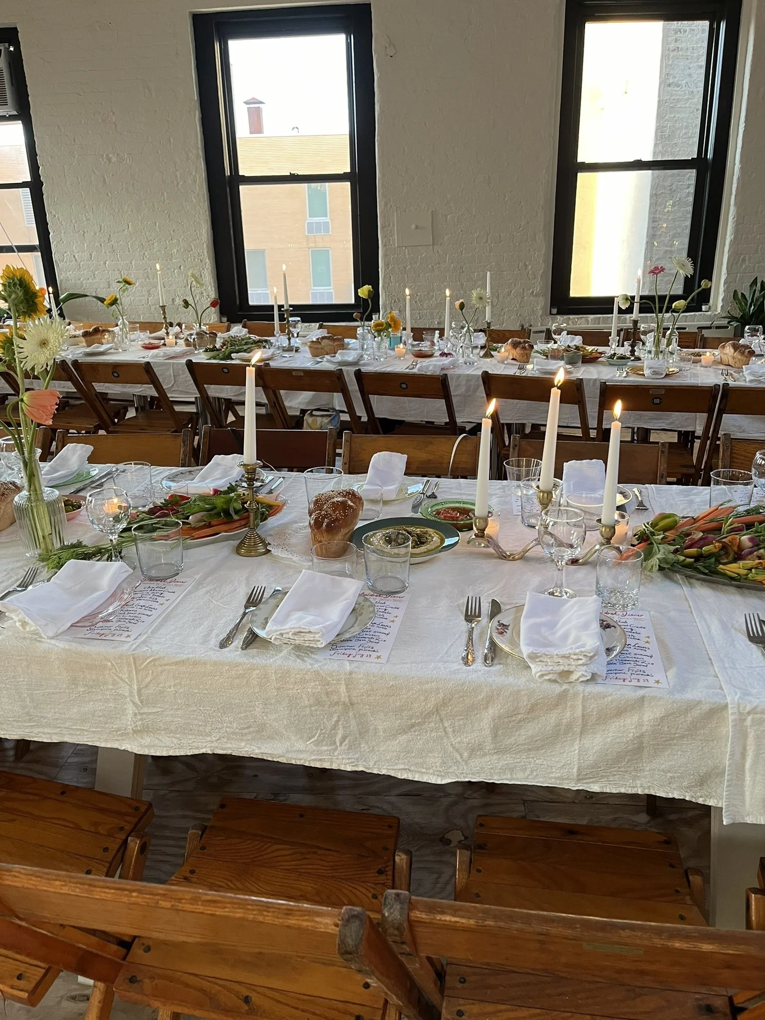Two rows of tables with vintage wooden folding chairs set for a Shabbos dinner with windows and city views in the background.