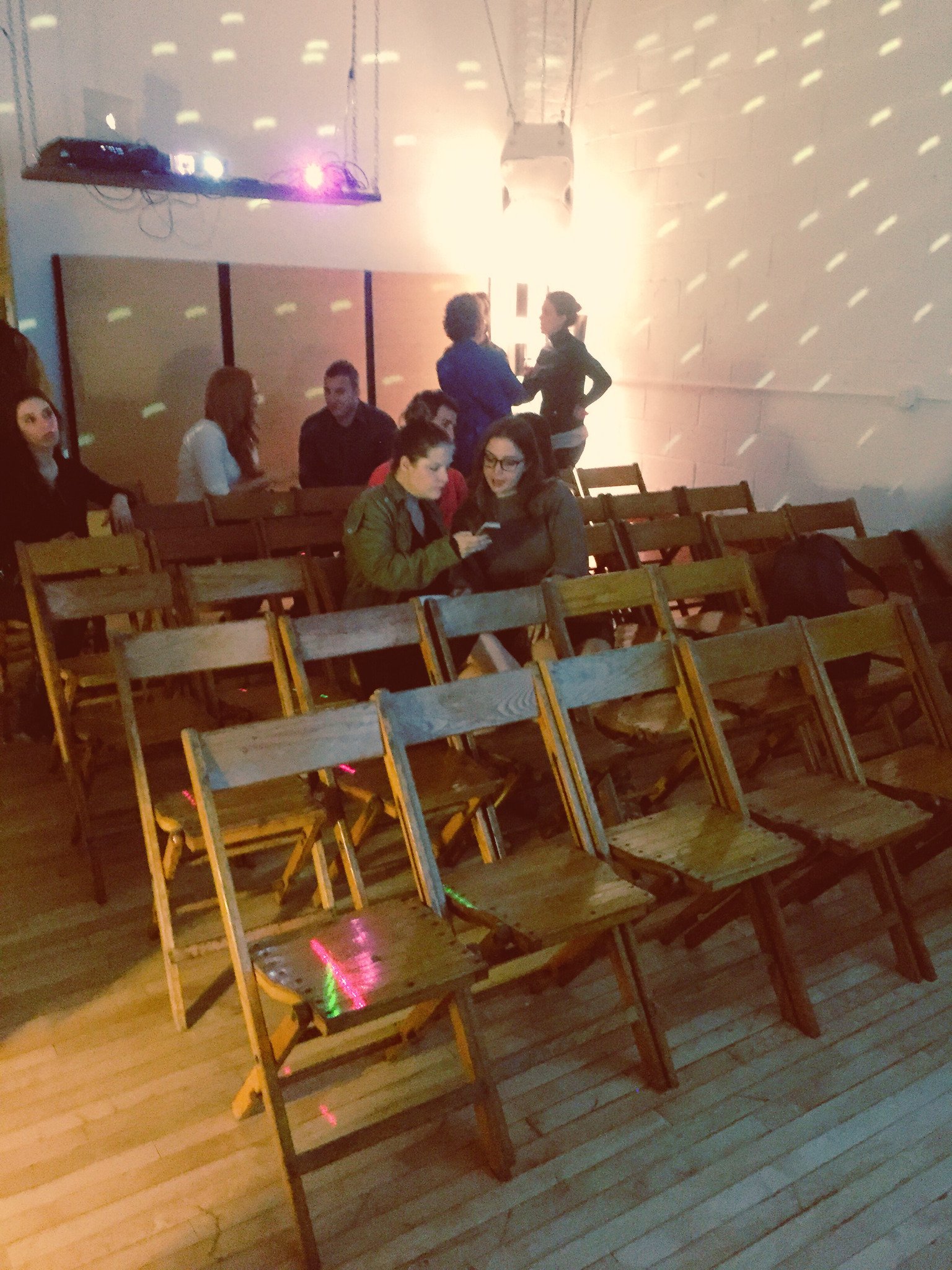 People socializing in a dimly lit room with wooden chairs, some seated at tables and others standing in small groups, illuminated by colorful lights and a projector casting patterns on the walls.